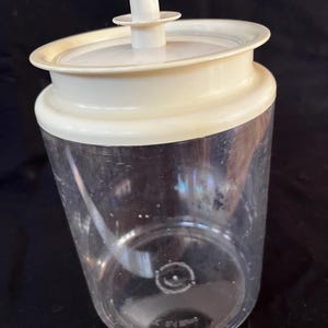 Op de afbeelding: Een doorzichtige plastic container met een wit deksel en een witte handgreep bovenop. De container is cilindrisch met een licht schuin ontwerp. Het deksel heeft een cirkelvorm met een verhoogde rand. De container is leeg.