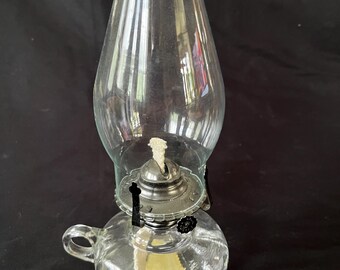 May include: A clear glass oil lamp with a round base and a handle. The lamp has a metal chimney and a wick.