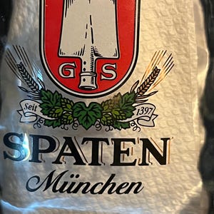 May include: A beer bottle label with the Spaten logo. The label has a red and white crest with a shovel design, the letters "G" and "S", and the words "SPATEN München". The label also includes the year "1397".