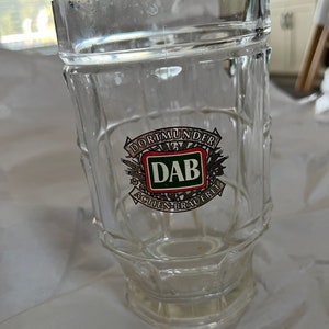 May include: A clear glass beer stein with a logo for Dortmunder DAB Aktien-Brauerei. The logo features a green shield with the letters DAB in white, surrounded by wheat stalks and a red banner.