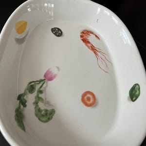 May include: White oval serving dish with painted illustrations of food items. The dish features a shrimp, a radish, a carrot slice, a black olive, a green vegetable, and a yellow egg. The dish has a handle on each side.