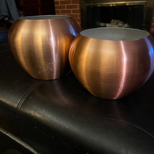 以下が含まれることがあります： 2つの丸い、金属製の、ローズゴールド色のプランター。プランターは黒い革の表面の上に置かれています