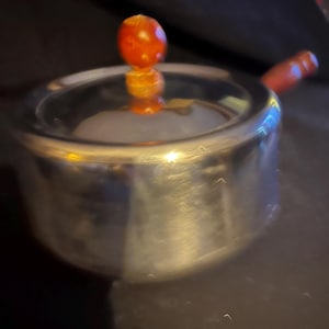 Pode incluir: Um açucareiro de prata com uma tampa de madeira e um botão vermelho. O pote tem uma superfície brilhante e reflexiva.