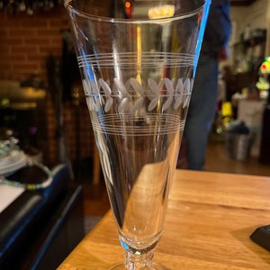 Op de afbeelding: Een helder glazen kelk met een lange, slanke steel en een brede, ronde voet. De kelk heeft een delicaat gegraveerd ontwerp van bladeren en lijnen rond het midden.
