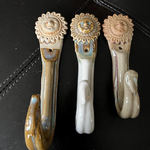 Puede incluir: Tres ganchos de cerámica con caras de sol. Los ganchos están esmaltados en tonos de marrón, blanco y azul. Cada gancho tiene un diseño de cara de sol diferente.