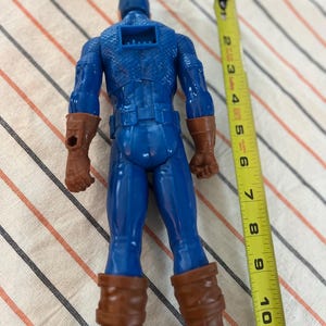 May include: A blue and red plastic action figure of Captain America, standing approximately 10 inches tall. The figure is wearing a blue and red suit with a red star on the chest. The figure is holding a shield in its right hand.