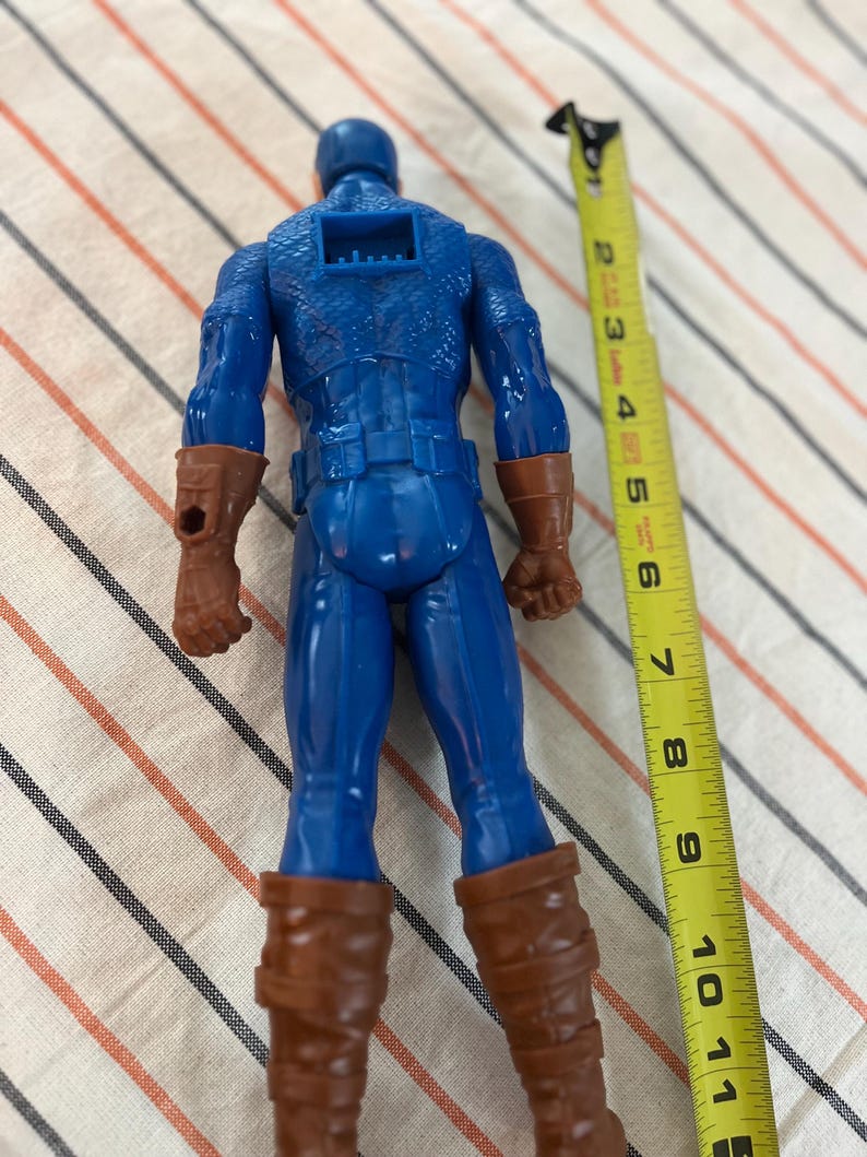May include: A blue and red plastic action figure of Captain America, standing approximately 10 inches tall. The figure is wearing a blue and red suit with a red star on the chest. The figure is holding a shield in its right hand.