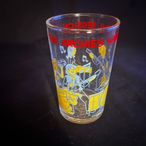 May include: A clear glass with a yellow and blue illustration of The Archies band playing music. The text "The Archies" and "Music Session" are printed in red on the glass.