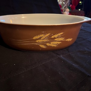Könnte beinhalten: Braune Pyrex-Backform mit einem Weizenmuster in Gold. Die Schüssel hat einen weißen Innenraum und eine braune Außenseite. Das Weizendesign befindet sich an der Seite der Schüssel. Die Schüssel hat zwei Griffe.