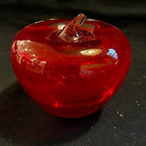 Może przedstawiać: Żywy, czerwony szklany jabłko. Ozdobny owoc ma łodygę i gładki, zaokrąglony kształt. Ten solidny, przyciągający wzrok element jest idealny do dekoracji wnętrz. Jabłko ma żywy odcień czerwieni.
