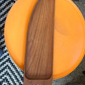 Puede incluir: Una bandeja de servir de madera con dos patas. La bandeja es marrón y tiene un acabado de madera natural. Está colocada encima de una bandeja redonda de color naranja brillante.