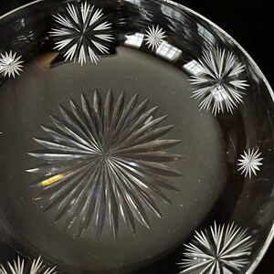 May include: A clear glass plate with a starburst pattern etched into the surface. The plate has a raised rim and a smooth, polished finish.