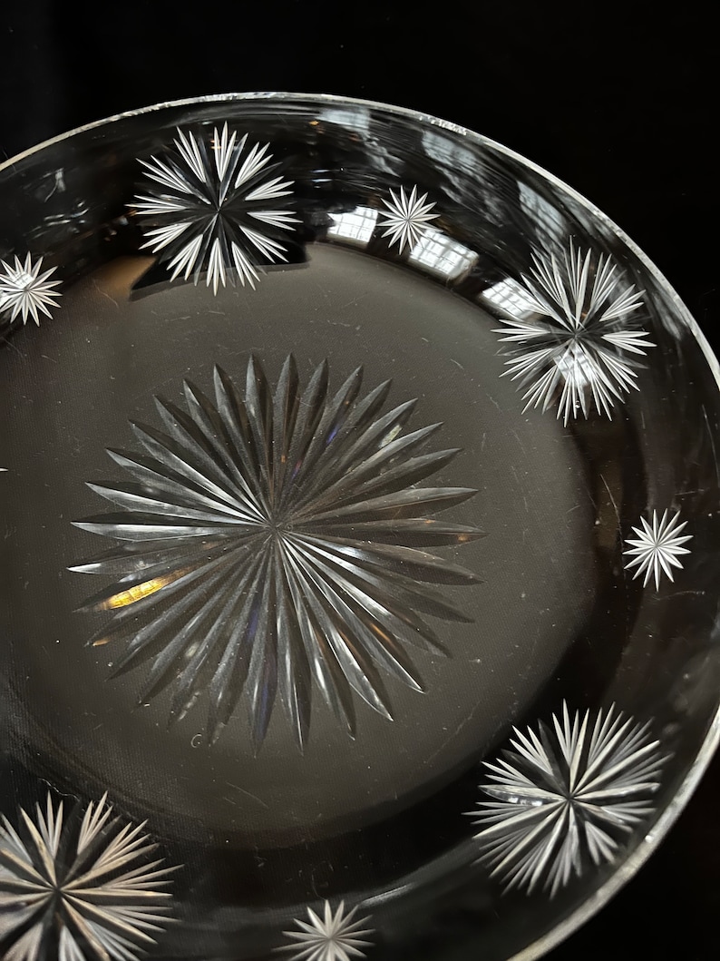 May include: A clear glass plate with a starburst pattern etched into the surface. The plate has a raised rim and a smooth, polished finish.