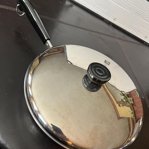 Puede incluir: Una sartén de acero inoxidable con mango y tapa negros. La tapa tiene un pomo negro en el centro. La sartén es redonda y refleja el entorno. El mango tiene un lazo para colgar.