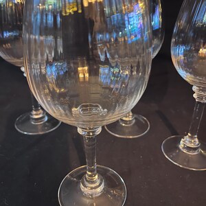 May include: Set of clear, ribbed wine glasses. The glasses have a classic design with a round bowl, a long stem, and a circular base. The glass reflects light, creating a shimmering effect. Ideal for serving wine at a dinner party.
