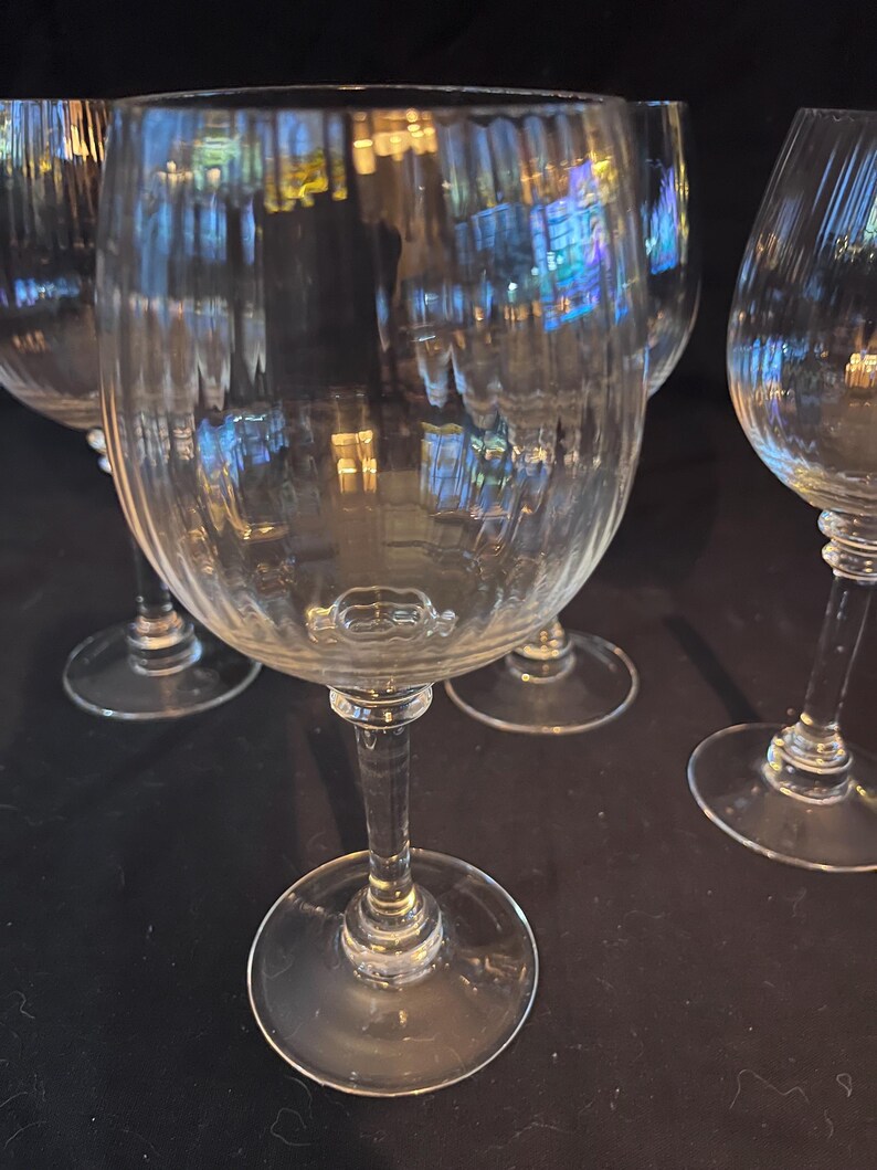May include: Set of clear, ribbed wine glasses. The glasses have a classic design with a round bowl, a long stem, and a circular base. The glass reflects light, creating a shimmering effect. Ideal for serving wine at a dinner party.