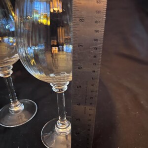 May include: Two clear wine glasses with textured bowls and long stems. A metal ruler is placed next to the glasses, showing measurements in inches. The glasses are on a dark surface.