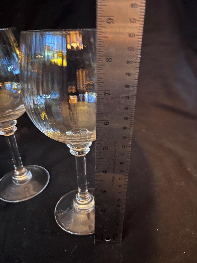 May include: Two clear wine glasses with textured bowls and long stems. A metal ruler is placed next to the glasses, showing measurements in inches. The glasses are on a dark surface.
