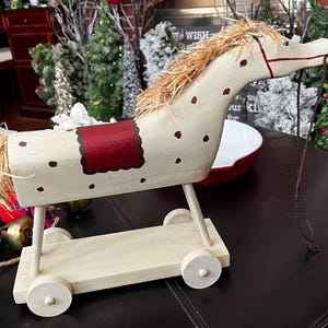 Op de afbeelding: Een crème geschilderd houten speelgoedpaard op wielen, met rode details zoals een zadel en stippen. Het heeft een manen en staart van stro en een bruine touwteugel. Een decoratief speelgoed in vintage stijl.