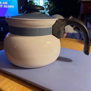 Könnte beinhalten: Eine weiße Teekanne aus Keramik mit einem blauen Deckel und einem schwarzen Henkel. Die Teekanne hat ein blaues Band um die Mitte.