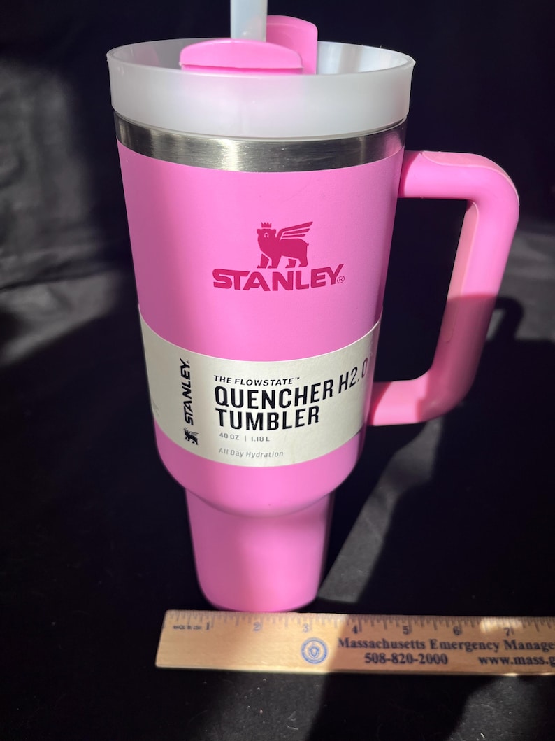 May include: A pink Stanley Quencher H2.0 tumbler with a handle, straw, and lid. The tumbler has the Stanley logo and text that reads "THE FLOWSTATE QUENCHER H2.0 TUMBLER 40 OZ | 1.18L All Day Hydration".