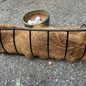 Puede incluir: Un macetero colgante de metal negro con un forro de coco marrón. El macetero está vacío y listo para plantas.