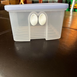 Könnte beinhalten: Ein durchsichtiger Plastikbehälter mit einem hellblauen Deckel. Der Behälter hat zwei weiße, runde Plastiklaschen an der Vorderseite.