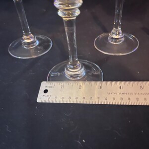 May include: Three clear stemmed glasses with round bases. A ruler shows the base diameter is approximately 3 inches. The stems have a decorative bulbous design. The glasses are on a dark surface.