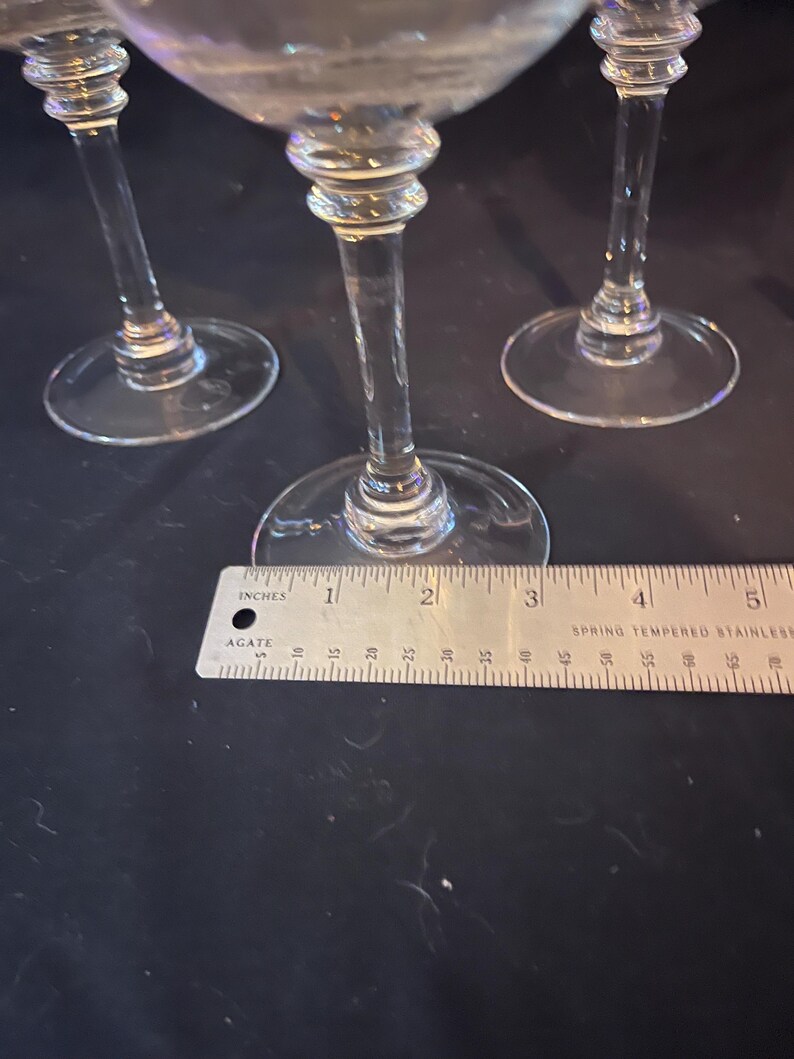 May include: Three clear stemmed glasses with round bases. A ruler shows the base diameter is approximately 3 inches. The stems have a decorative bulbous design. The glasses are on a dark surface.