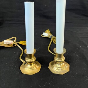 Puede incluir: Dos lámparas de velas eléctricas vintage. Cada lámpara presenta una vela cilíndrica blanca sobre una base hexagonal de color latón. Las lámparas están conectadas a una fuente de alimentación mediante un cable amarillo.
