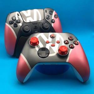 May include: Two custom video game controllers featuring a Volkswagen logo design. The controllers are black and grey with pink accents and red buttons. The top controller is a PlayStation 5 controller and the bottom controller is an Xbox controller.