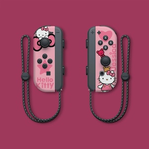 May include: A pink and black Nintendo Switch Joy-Con controller set with a Hello Kitty design. The left Joy-Con features a Hello Kitty graphic with the text "Hello Kitty" and the right Joy-Con features a Hello Kitty graphic with the text "Jessica".