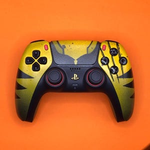 May include: A black and yellow PlayStation 5 DualSense controller with a custom design. The controller has a yellow and black tiger stripe pattern with a black and yellow abstract design on the touchpad.