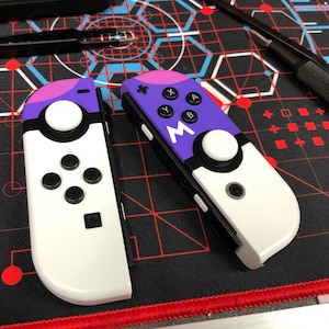 May include: Two custom-designed video game controllers. The controllers feature a white base with purple and pink accents. One controller has black buttons and a white joystick. The other controller has black buttons, a white joystick, and the letter "M".