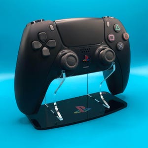 May include: A black PlayStation 5 DualSense controller on a black acrylic stand with the PlayStation logo. The stand has two clear acrylic supports.