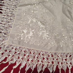 May include: A square off-white shawl featuring intricate tone-on-tone floral embroidery across the fabric surface. The edges are finished with a wide, decorative macramé border that transitions into long, flowing fringe tassels along the sides.