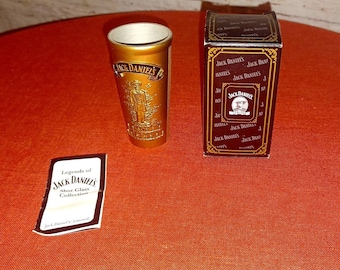 Puede incluir: Un vaso de chupito dorado con el logotipo de "Jack Daniel's" y una representación de un hombre. Junto a él, una caja con el logotipo y el texto de "Jack Daniel's". También hay una pequeña tarjeta con el texto "Legends of Jack Daniel's Shot Glass Collection".