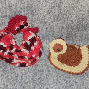 May include: Two crocheted items, one shaped like a rooster in shades of red, white, and dark maroon, and one shaped like a small bird in shades of tan and brown. Both items feature small black circular accents for eyes and textured yarn construction.