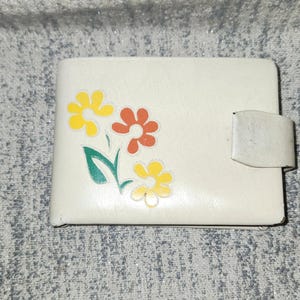 May include: Cream-colored wallet with a floral design. The wallet features a clasp closure and is decorated with yellow, orange, and yellow flowers with green stems and leaves. The wallet is rectangular in shape.