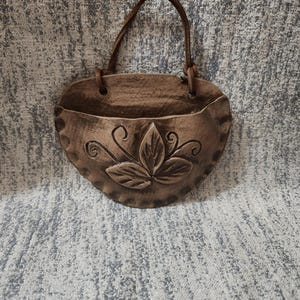 May include: A semi-circular brown ceramic wall pocket featuring a textured surface with a central embossed leaf motif and scalloped edges. The piece includes a brown leather strap attached through two circular holes at the top for hanging.