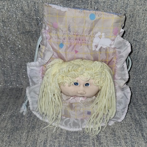 May include: A drawstring bag featuring a doll head with blonde yarn hair and blue eyes. The bag is made of a light pink fabric with a yellow grid pattern, small blue and pink geometric shapes, and a ruffled white trim along the edges.