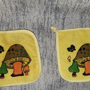 May include: A pair of square yellow pot holders featuring a central graphic of a large brown mushroom with a yellow underside, flanked by smaller green and orange mushrooms and a black butterfly, all finished with a yellow fabric border.