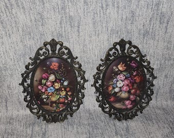 May include: Two vintage oval wall plaques with ornate bronze-colored frames. Each plaque features a floral still life painting with a variety of colorful flowers in a vase. The background is a dark, muted tone.