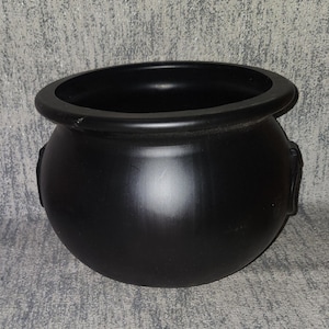 May include: A matte black cauldron with a rounded body, a wide circular opening, and a thick rim. The vessel features two small, integrated side handles and a smooth, uniform finish across its entire exterior surface, presenting a deep black color.
