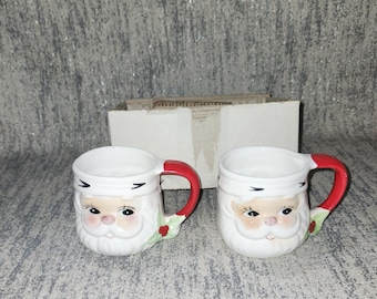 May include: Two white ceramic mugs with Santa Claus faces. Each mug has a red handle and a green holly accent. The Santa faces feature rosy cheeks, a white beard, and a black mustache.