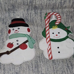 May include: Two fabric snowman ornaments. The left snowman wears a black hat with a red band, a green and white striped scarf, and holds a brown broom. The right snowman wears a green hat with holly and a green scarf, holding a red and white candy cane.