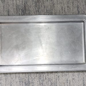 May include: A rectangular silver-toned metal serving tray with a smooth, flat base and a raised border. The tray features two ornate, black metal handles attached to the short sides, each displaying a detailed, symmetrical scrollwork design.