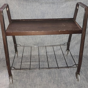 May include: A brown rolling utility cart featuring a solid top shelf with a wood grain finish and a lower shelf constructed from thin black metal wire. The frame is made of brown metal with two side handles and four small wheels at the base.
