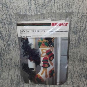 May include: A Christmas stocking kit in a clear plastic package. The kit includes a needlepoint stocking with a design of bells and holly. The package has the text "SANTA STOCKING" and the brand name "BERNAT".