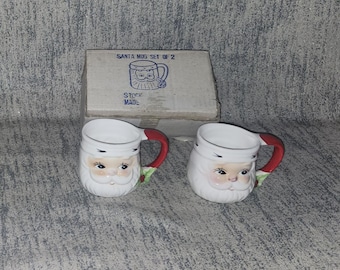 May include: Two ceramic Santa mugs with white faces, red and green handles, and a matching box. The mugs have a Santa Claus design with rosy cheeks and a white beard. The box reads "SANTA MUG SET OF 2".